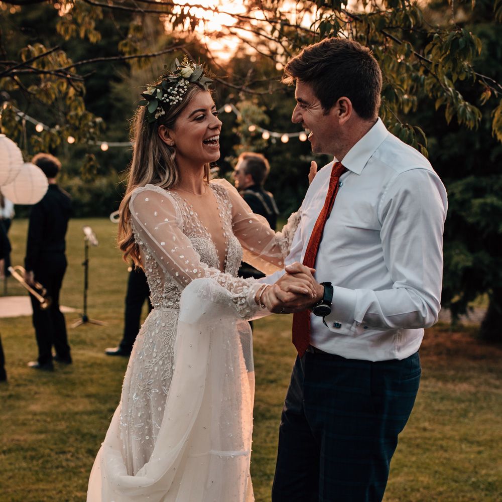 Bride in long sleeve v neck wedding dress and flower crown dances during golden hour with groom in white shirt a red tie at tipi wedding reception