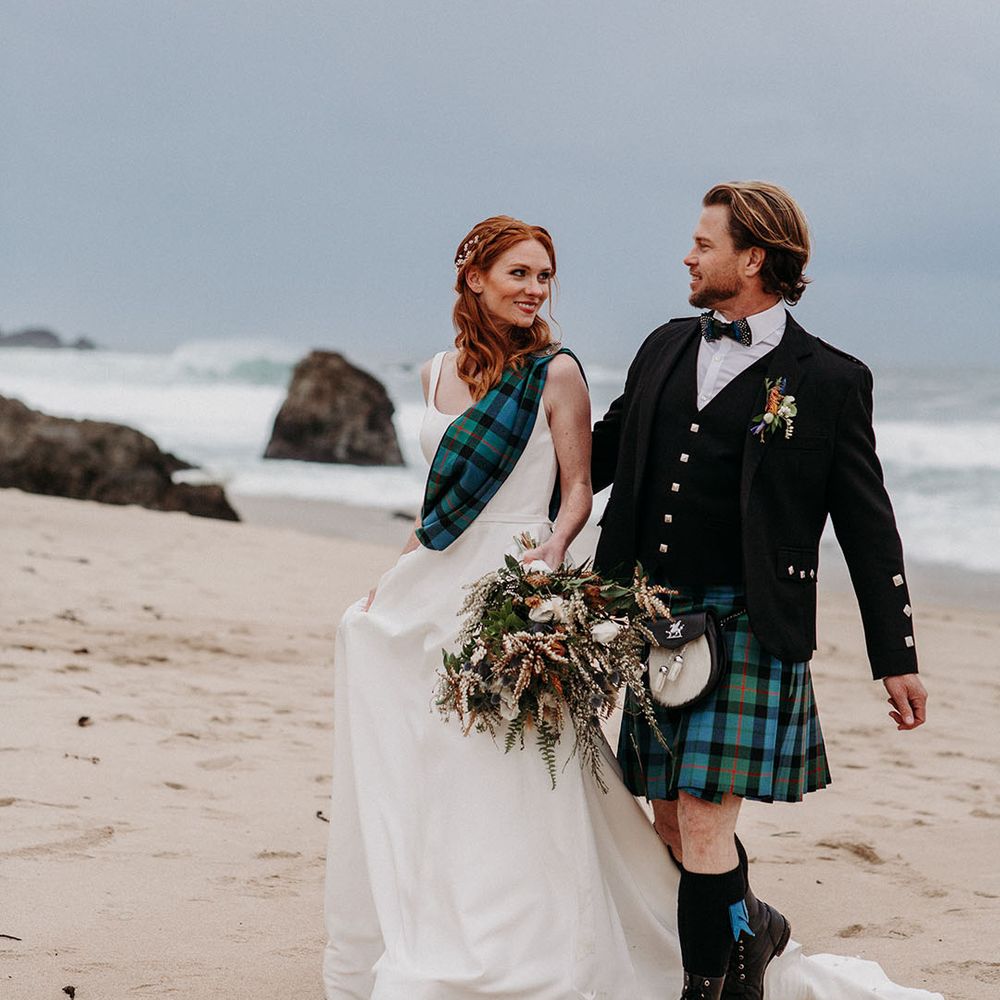 Bride in a princess wedding dress with tartan sash and groom in a kilt walking along the beach at Big Sur, California 
