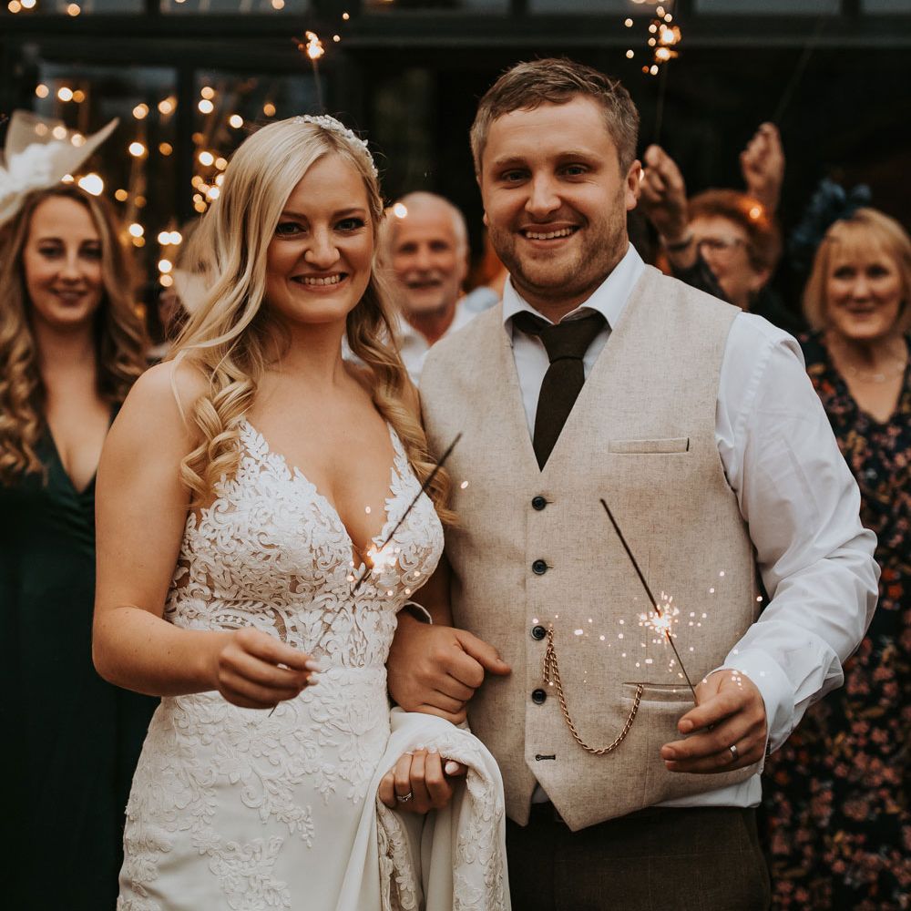 Bride in lace wedding dress holding train stands with groom in white shirt and beige waistcoat both holding sparklers at Hidden River Barn Wedding