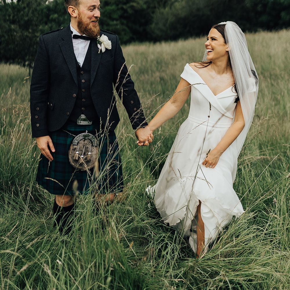 Luxury micro wedding with groom in a tartan kilt holding hands with the bride in an off the shoulder Suzanne Neville wedding dress