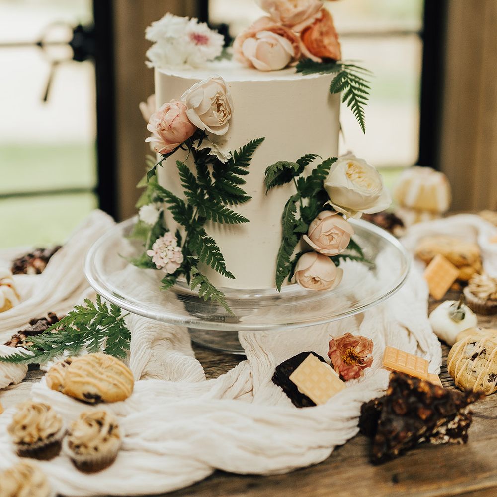 Single tier buttercream wedding cake on a glass cake stand decorated with flowers and fern leafs