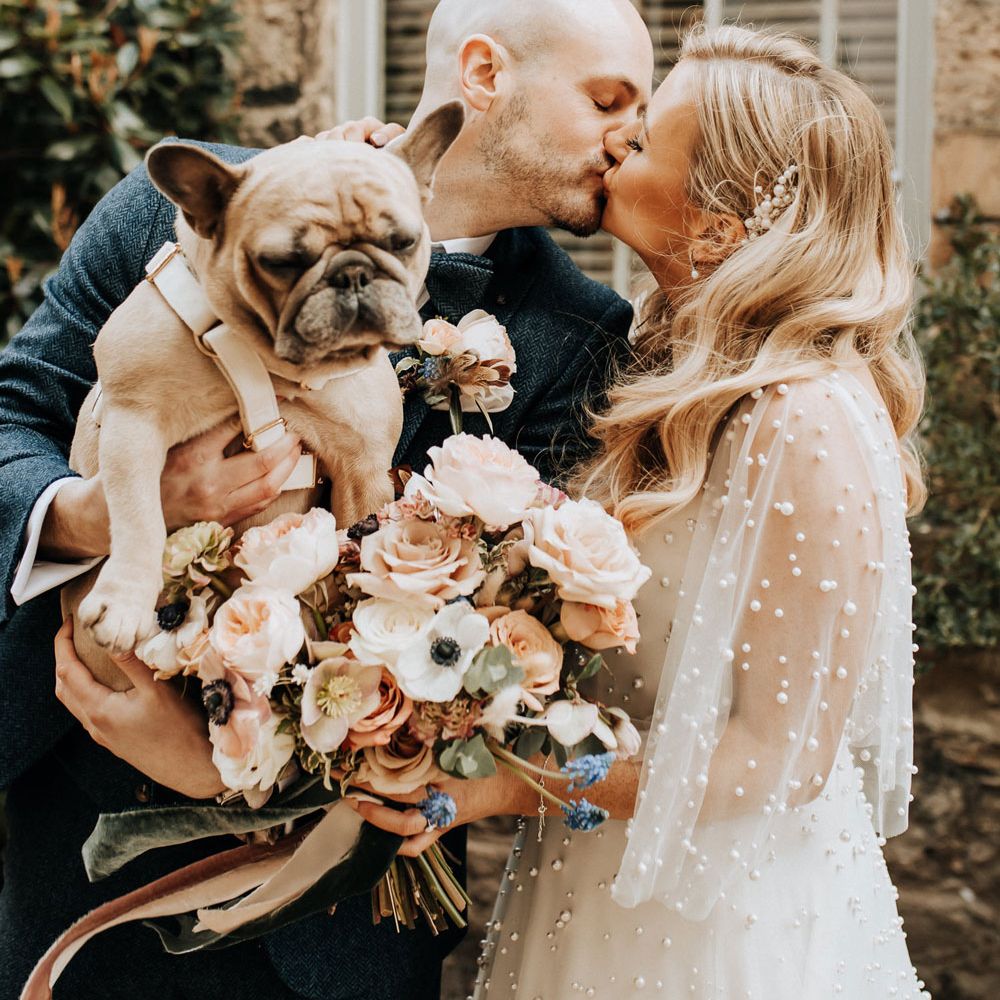 Bride in pearl wedding dress with sleeves kisses groom in navy tweed wedding suit holding French bulldog 