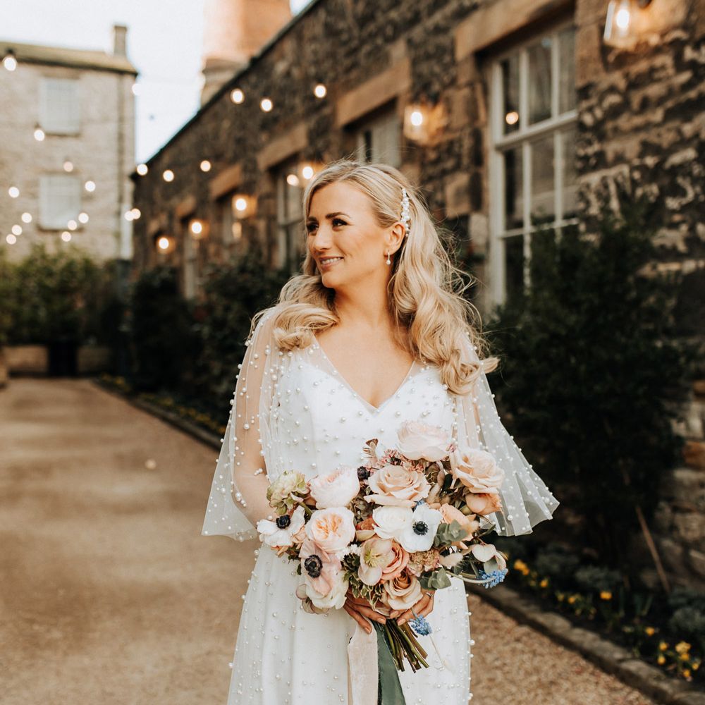 Bride with white short sleeve wedding dress with pearls stands holding mixed pastel wedding bouquet with roses and anemones