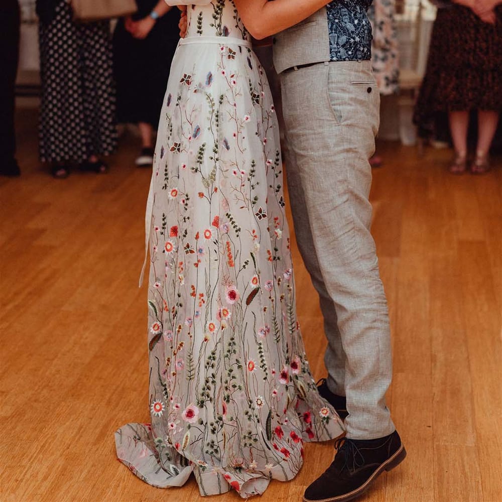 Bride in floral embroidered wedding dress dances with groom in grey and blue patterned waistcoat 
