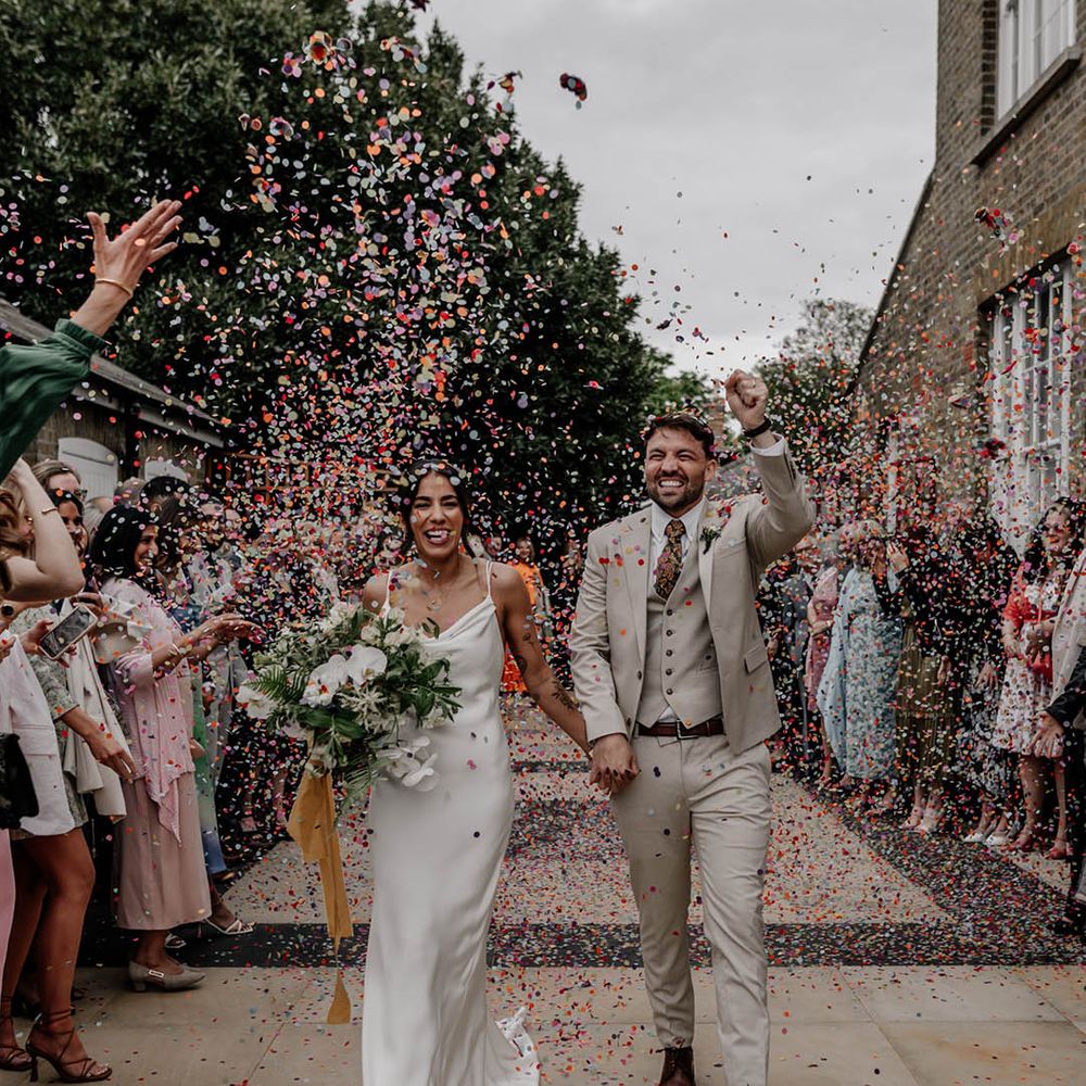 Colourful confetti photograph with bride in a satin wedding dress and groom in a beige three piece suit 