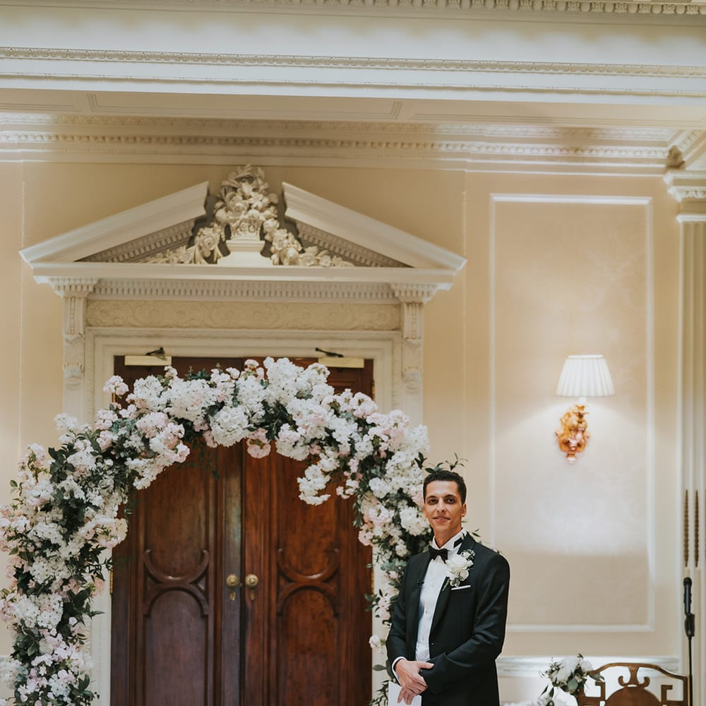 Pink and white wedding flower arch with groom in black tie waiting for the bride 