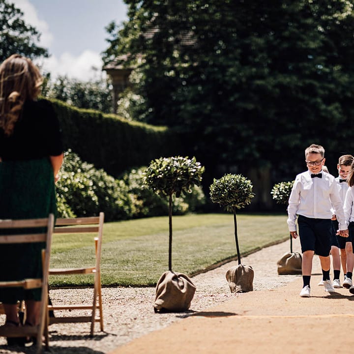 Page boys in black shorts, white shirt and black bow ties walks together followed by two flower girls in pink dresses and white flower crowns