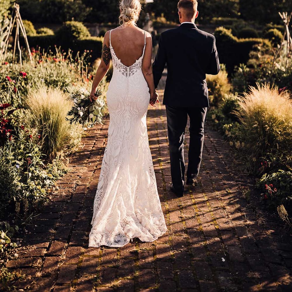 Bride in scoop back lace wedding dress with button details walks in the gardens holding hands with the groom in a blue tuxedo 