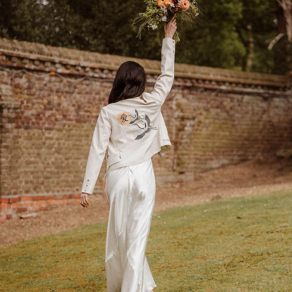 Bride in a satin slip wedding dress and white jacket with ancient Japanese motif holding her bouquet in the air at Gorse Hill