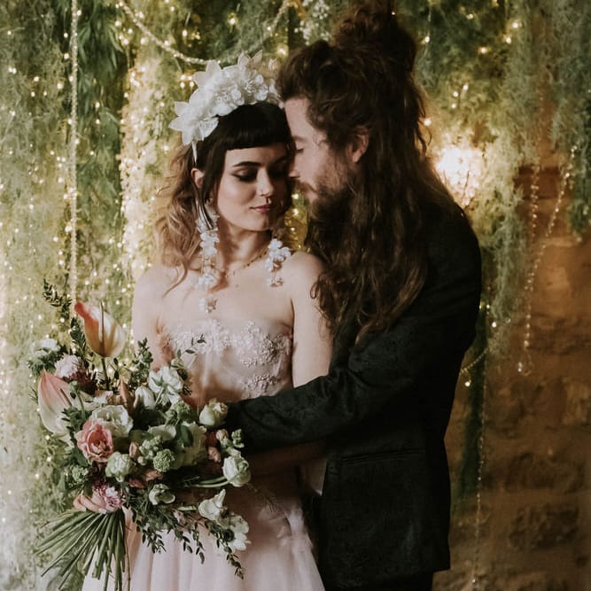 Groom in a black suit embracing his bride in a pink wedding dress as she holds a spring flower bouquet in front of a rustic twig, foliage and fairy light backdrop 