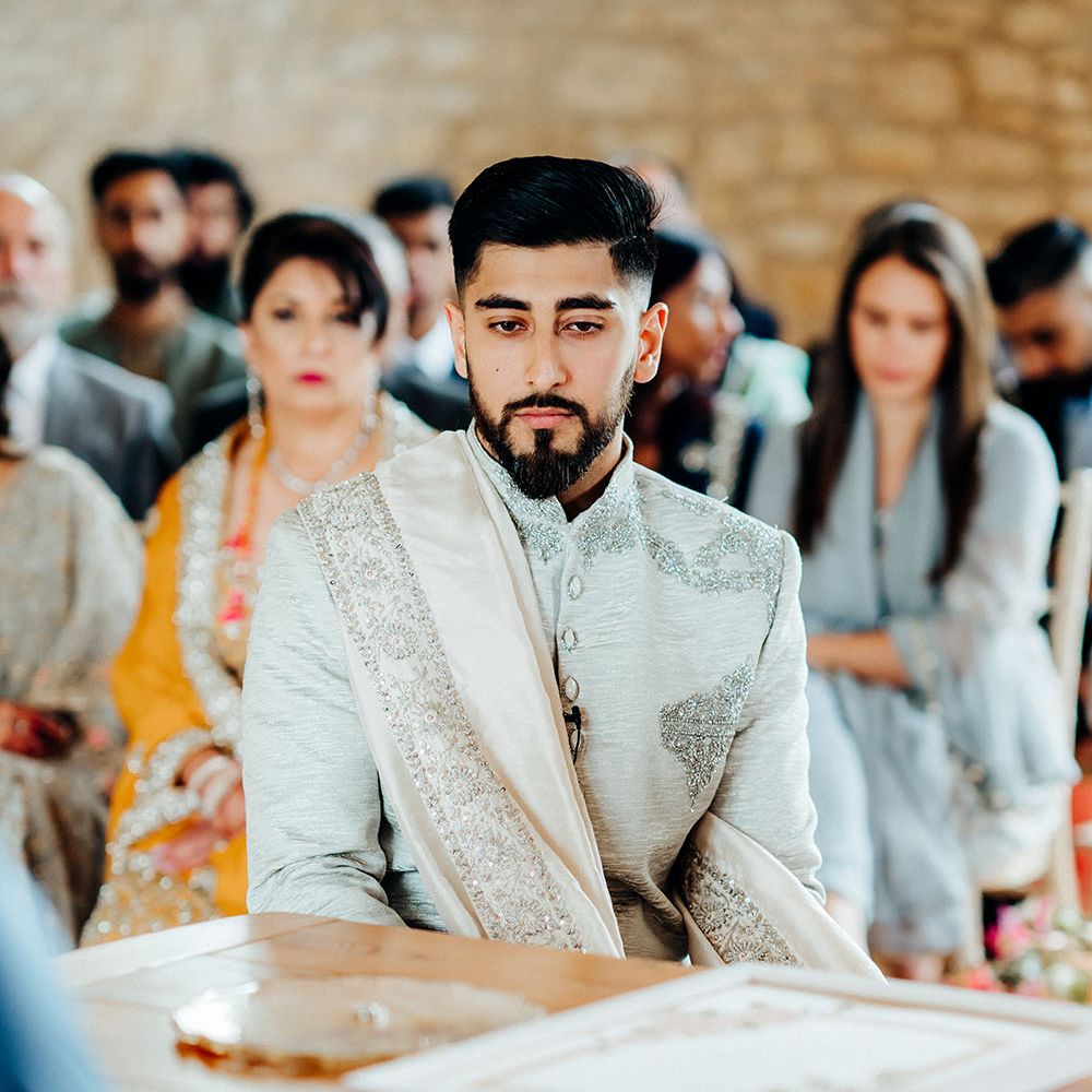 Groom wears light grey sherwani with embellishment whilst sat during wedding ceremony