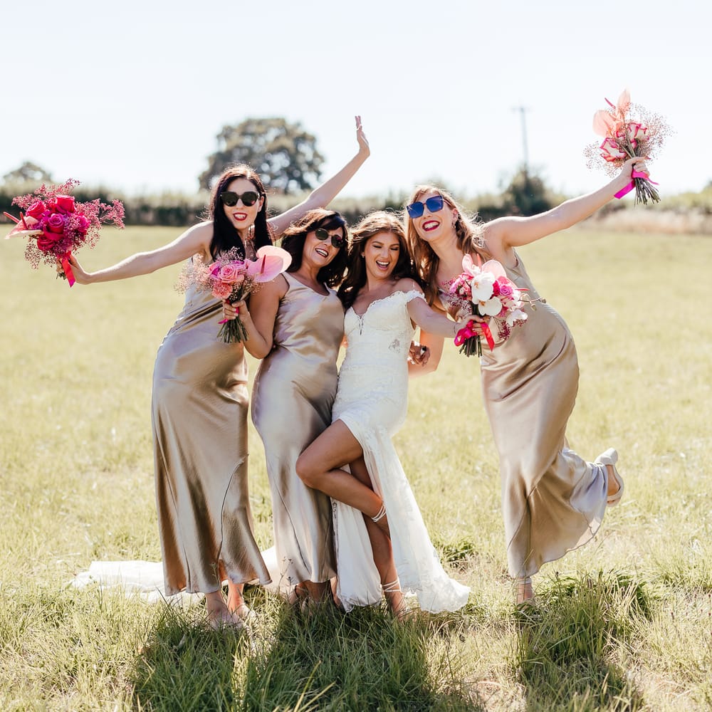 Bridesmaids wear sunglasses and hold out bright floral bouquets whilst wearing champagne bridesmaid dresses outdoors 