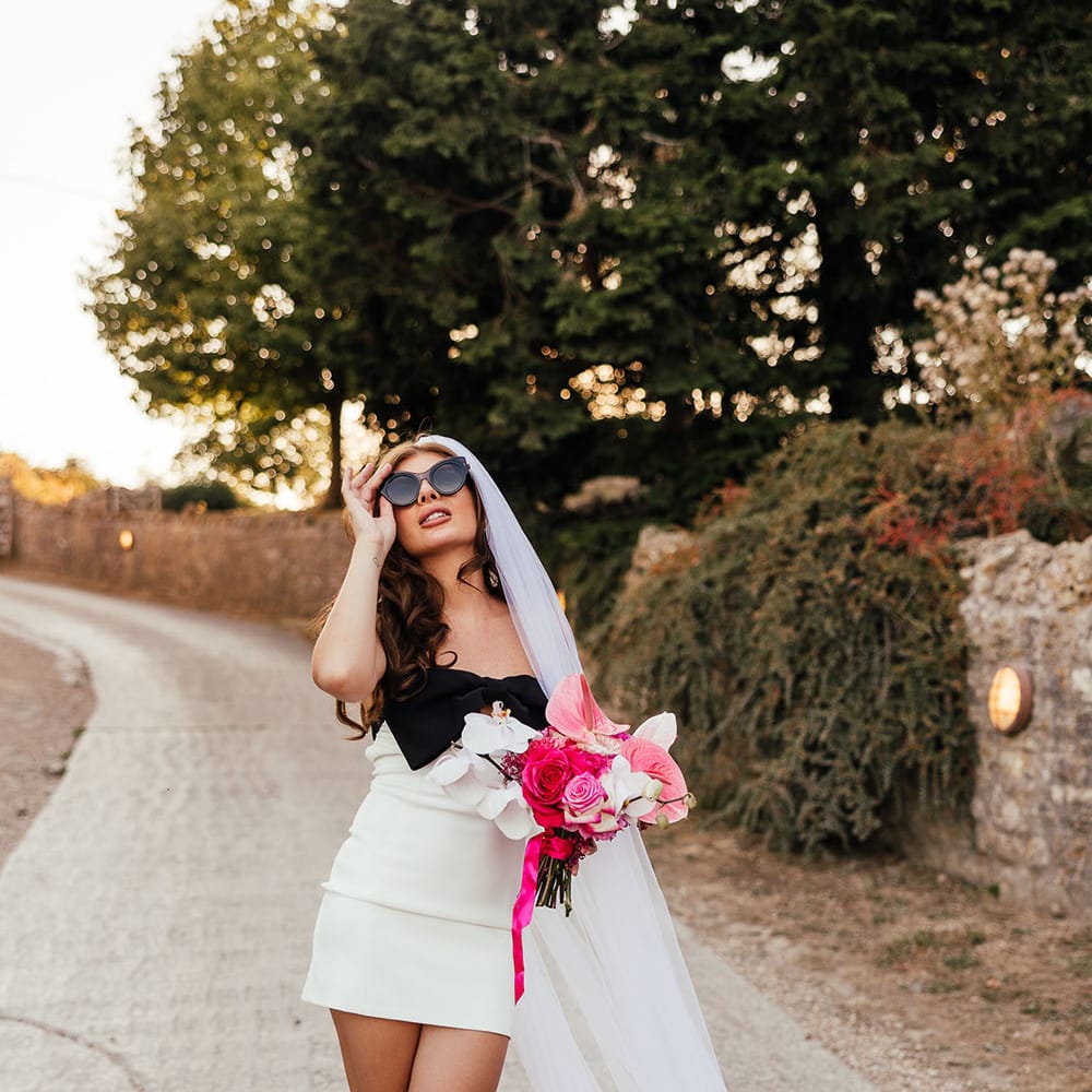 Bride wears white mini dress with black bow to front and cathedral veil whilst holding bright pink floral bouquet  