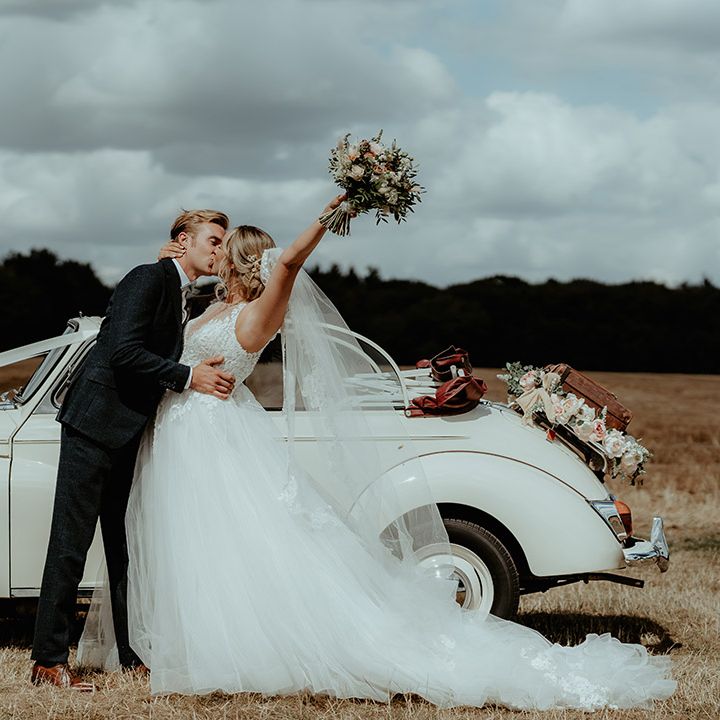 Cream vintage wedding car with white ribbon and flower decorations and bride and groom sharing a kiss 