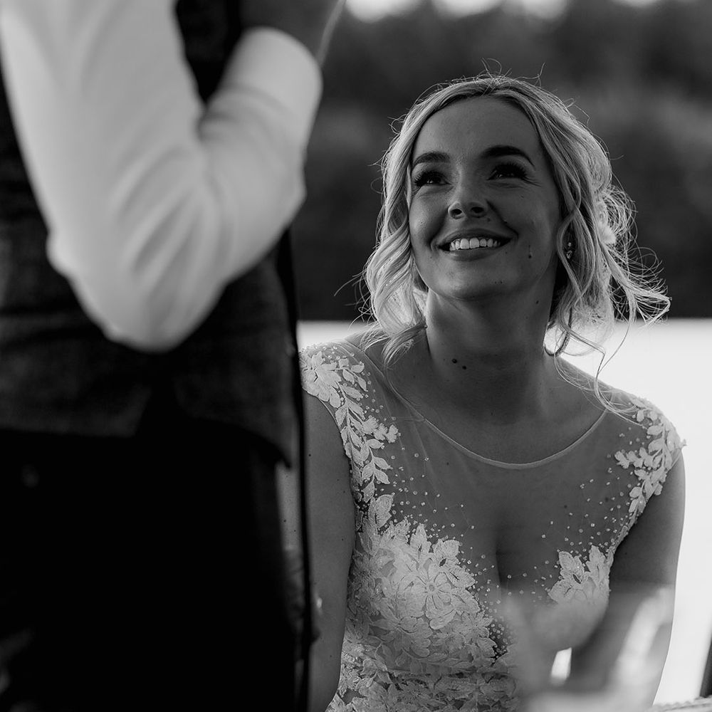 Bride in an embellished wedding dress with and illusion style neckline smiles up at the groom lovingly as he delivers his speech