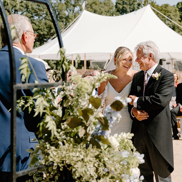 Bride stands beside her father who wears traditional morning suit during outdoor wedding in the summer