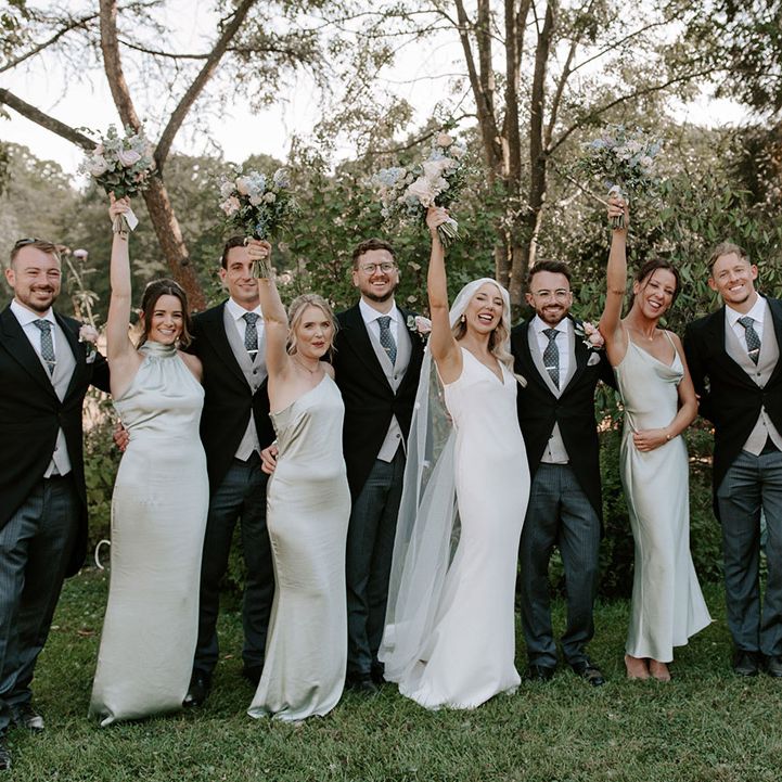 Bride and groom pose with their wedding party with bridesmaids in sage green dresses and groomsmen in three piece morning suits 