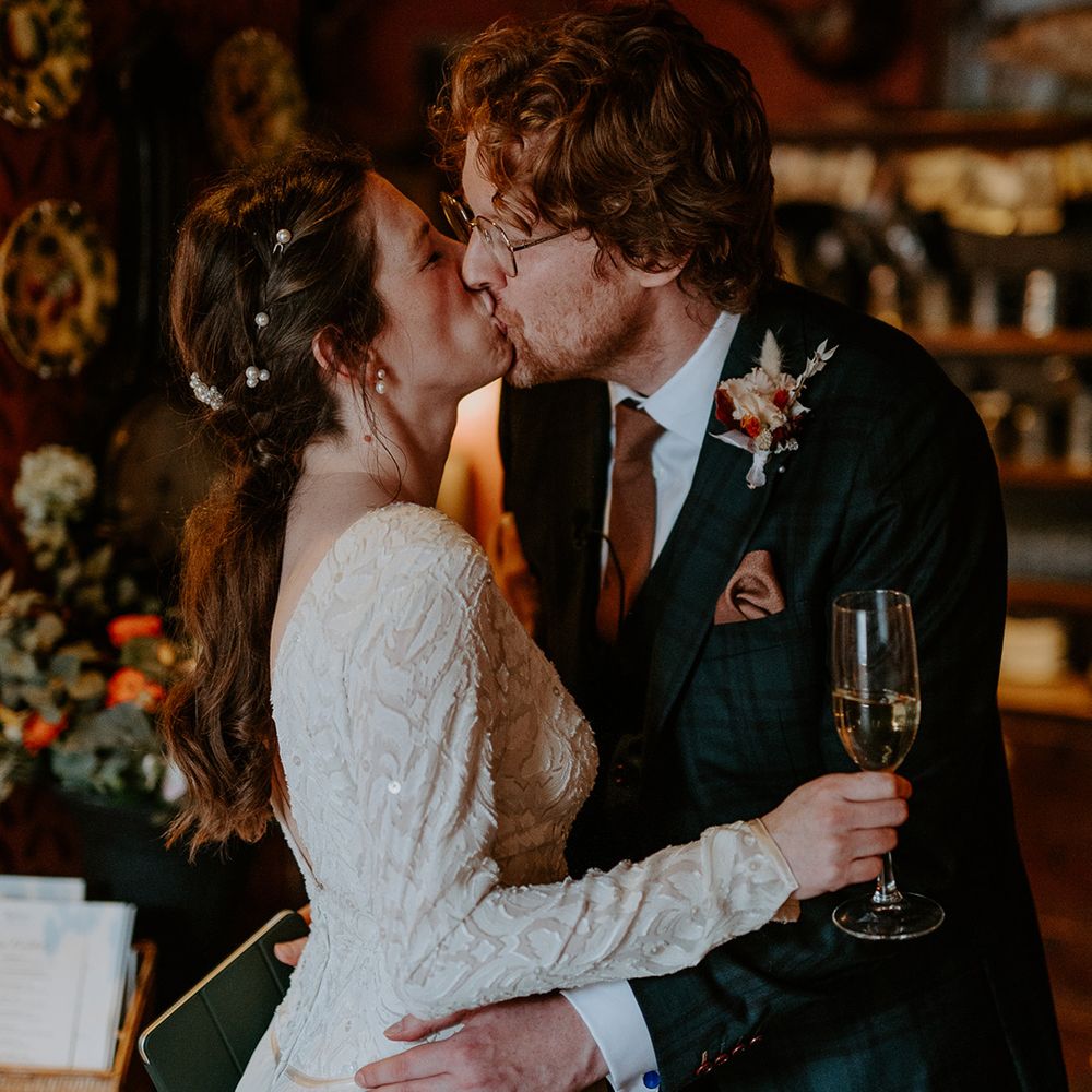 Bride in long sleeve lace vintage wedding dress, pearl earrings and pearl hair accessories kissing groom in dark suit with orange tie and autumnal boutonniere 