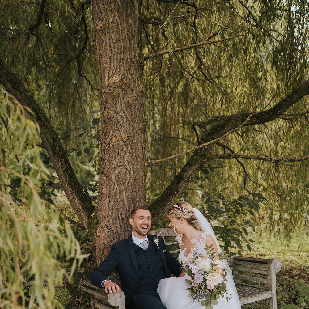 The bride in an illusion lace and tulle wedding dress with sparkly silver shoes sits on a wooden bench laughing with the groom in a navy suit 