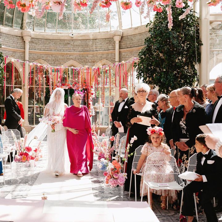 Mother of the bride in bright pink wedding outfit walking the bride down the aisle 
