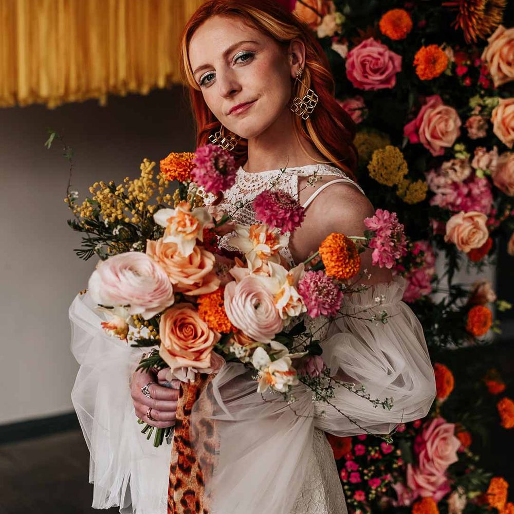 Bride in 70s inspired boatneck short lace wedding dress with detachable tulle puff sleeves and platform gold wedding heels holding large colourful mixed flower bridal bouquet tied with leopard print ribbon 