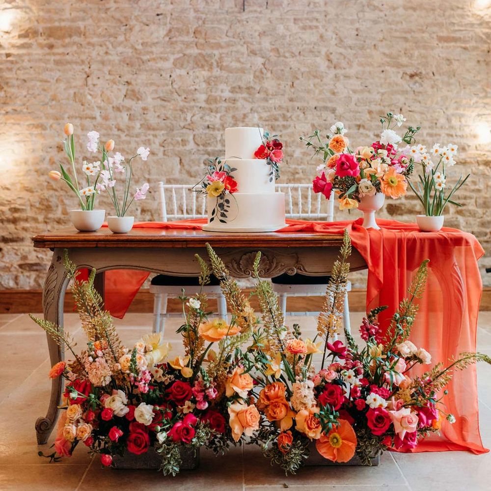 Merriscourt wedding venue reception room with wooden table with orange table runner and mixed seasonal flower arrangements and misshapen three tiered wedding cake with white icing and seasonal flower and foliage cake toppers 