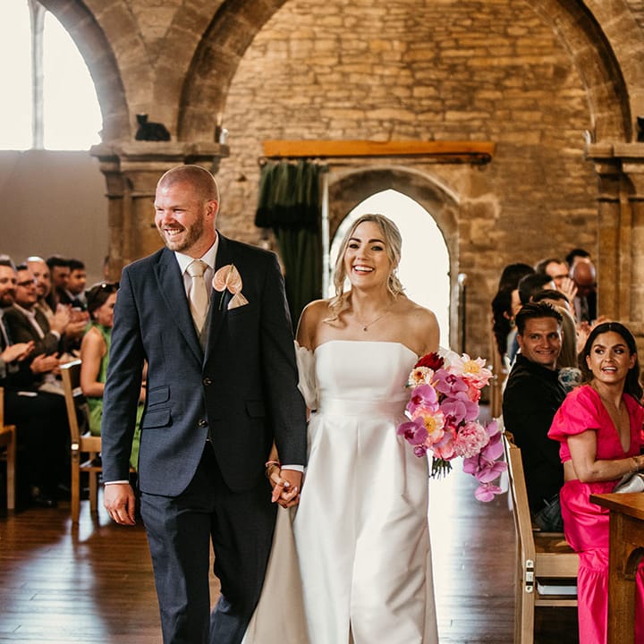 Bride in off the shoulder Jesus Peiro wedding dress with off the shoulder detail with the groom in a navy suit exiting from their church ceremony 