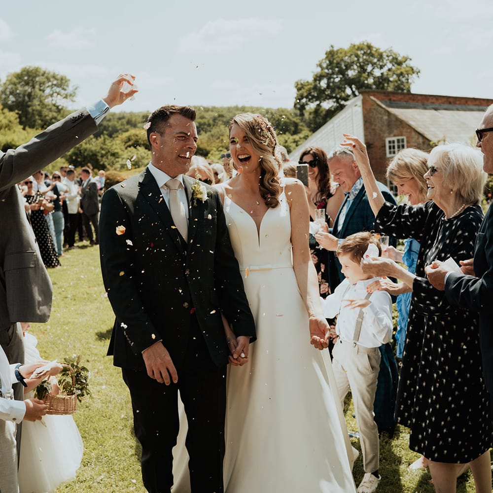 Bride in traditional white wedding dress with groom in suit with pale pink tie walking out of the ceremony to confetti 
