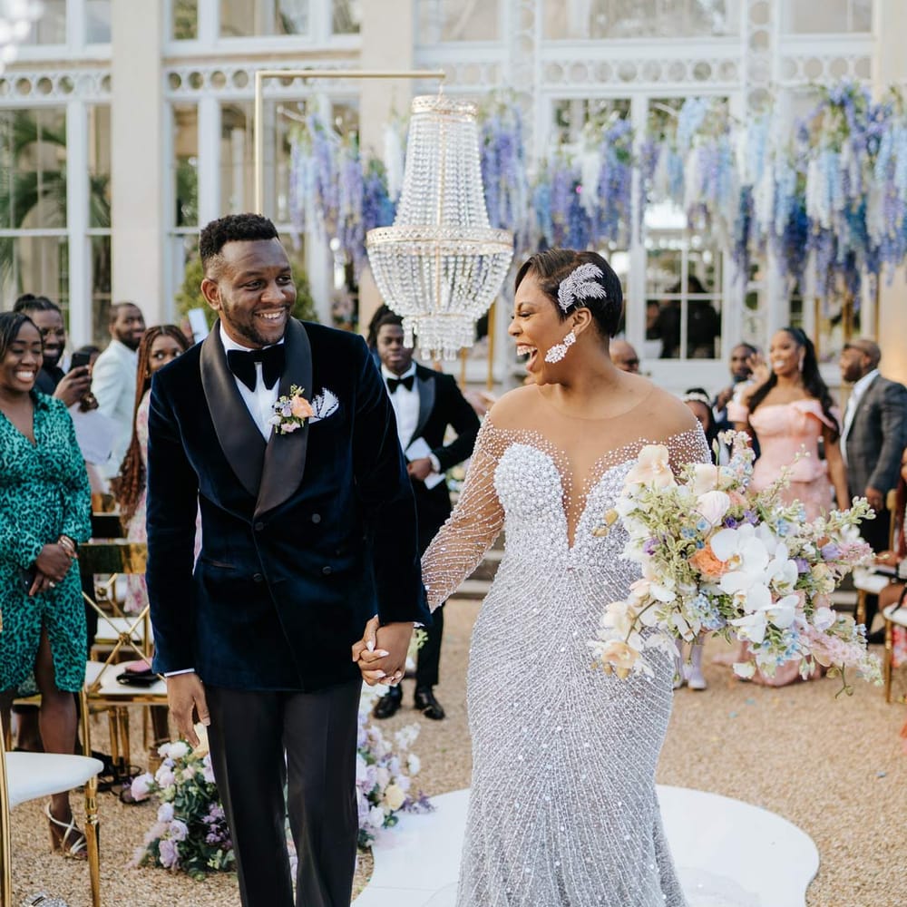 Bride in illusion v neck sheer off shoulder long sleeve pearl embellished wedding gown and groom in velvet midnight blue double breasted groom’s blazer, velvet bowtie and peach garden rose boutonniere walking down the aisle at Syon Park vow renewal 