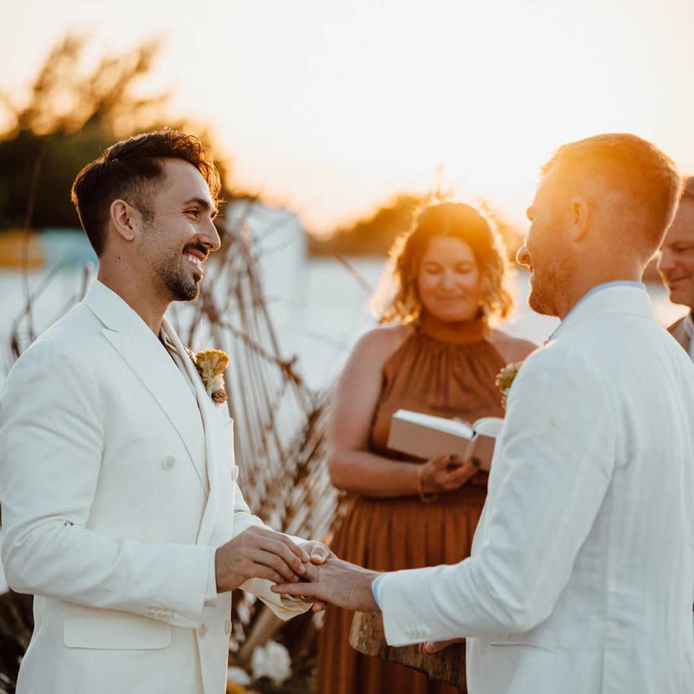 Golden hour wedding portrait with two grooms in white wedding suits exchanging vows on the beach 