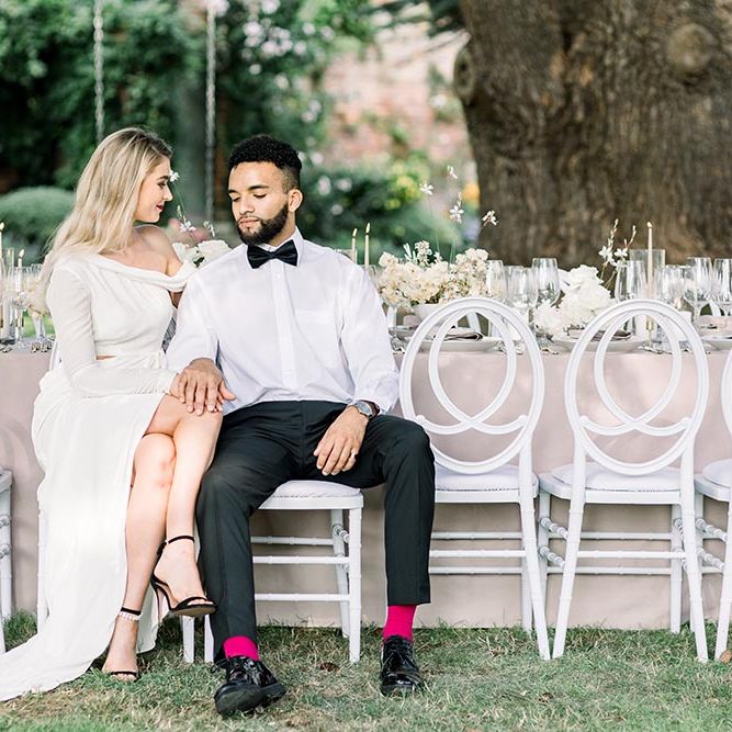 Sophisticated bride and groom sitting at their outdoor wedding reception on white chairs 