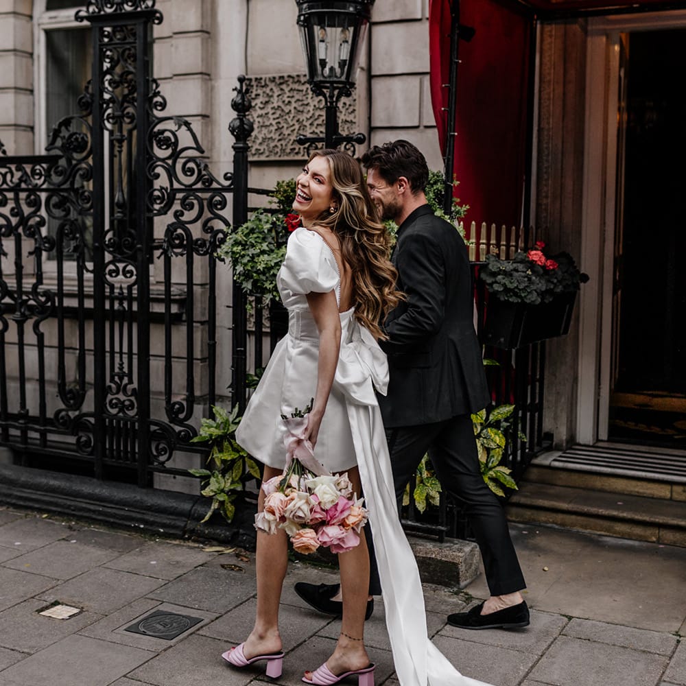 Portrait of a bride in a short wedding dress with long trailing bow back detail walking through Mayfair with her groom 
