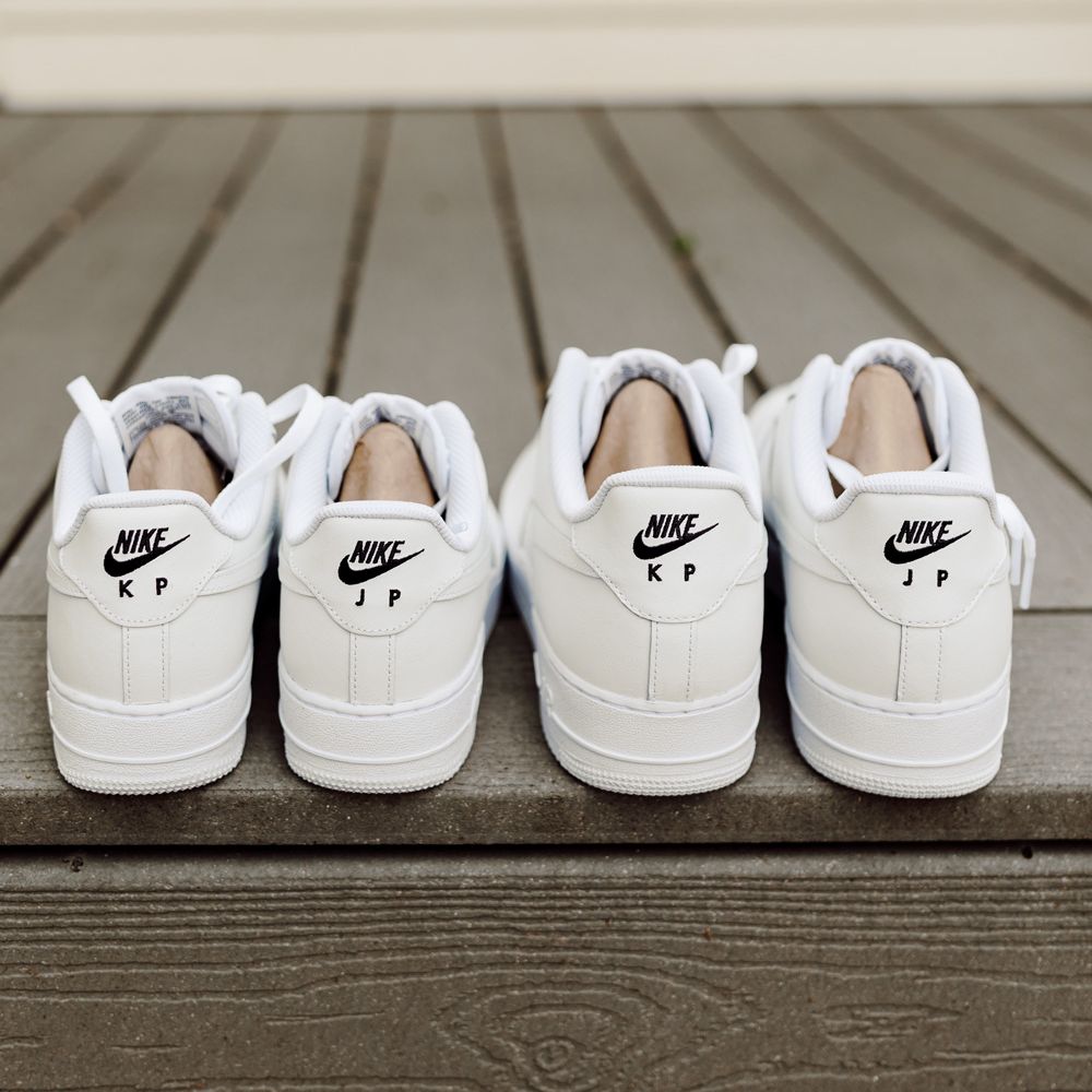 Two pairs of white Nike Air Force 1 trainers with black Nike logo and black embroidered initials on the back resting on wooden porch at backyard wedding