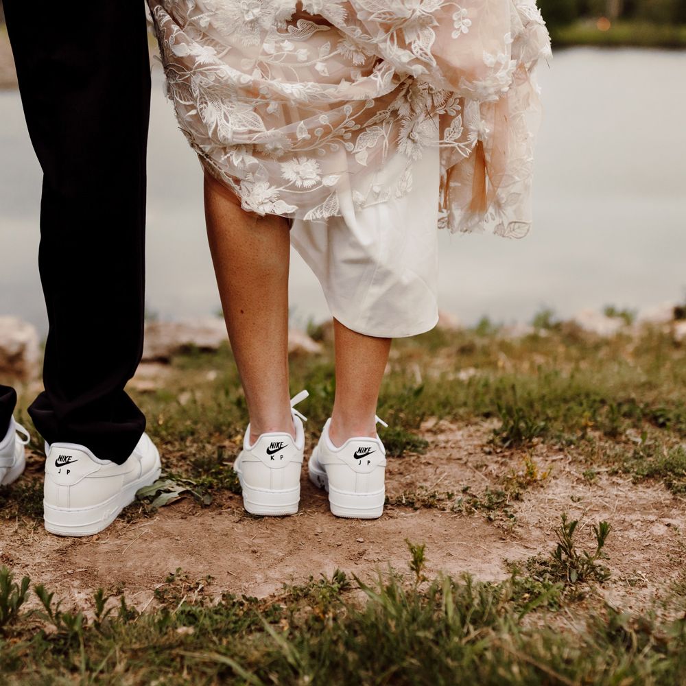 Groom in black trousers and bride holding up white embroidered Made With Love wedding dress wear matching personalised white Nike Air Force 1 trainers as they stand next to lake after backyard wedding