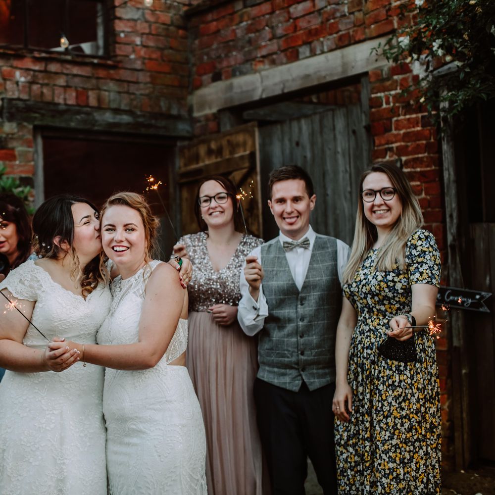 Bride kisses her bride on the cheek as they are surrounded by wedding guests holding sparklers outdoors