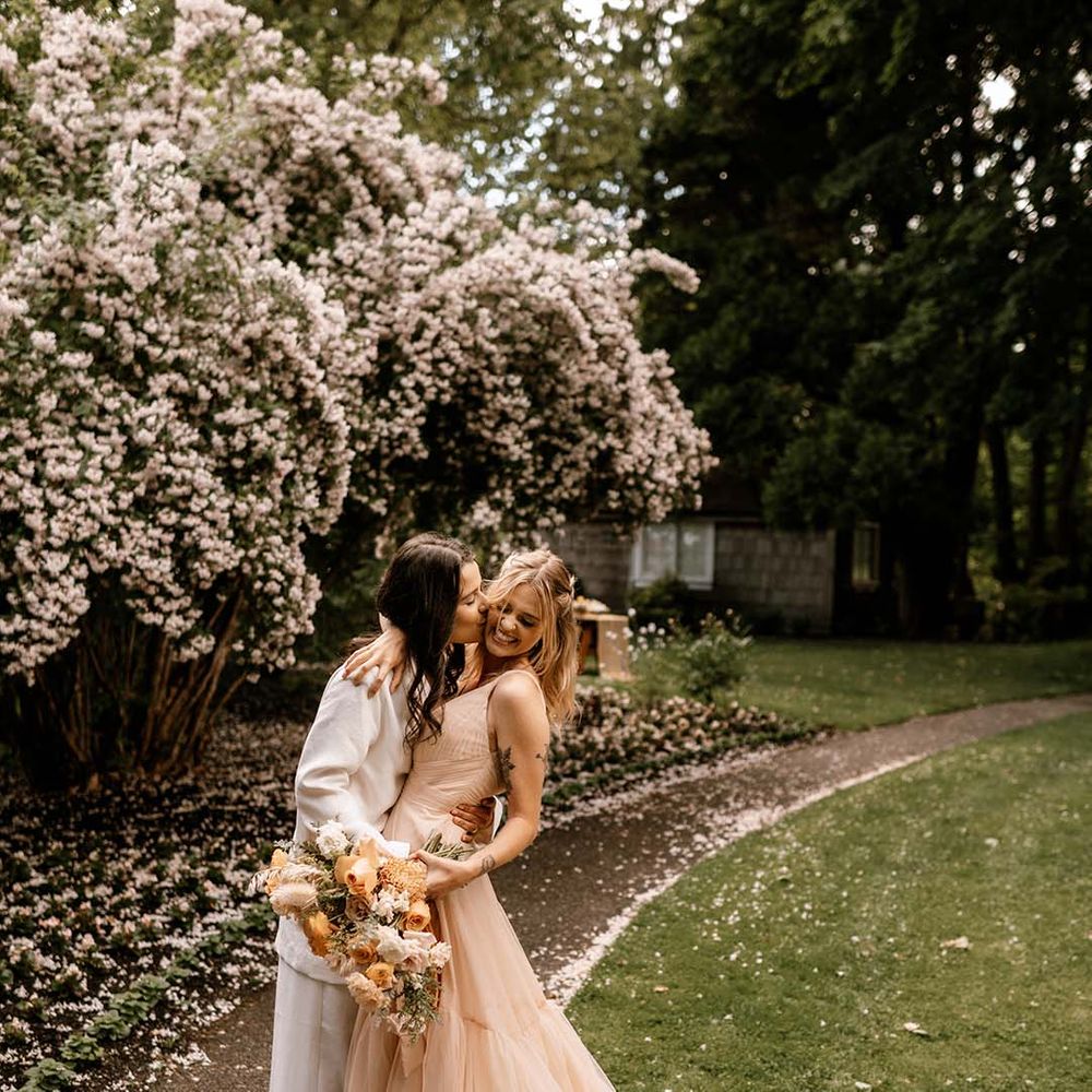 Bride in a white suit kissing her brides cheek in a blush pink wedding dress as she holds a yellow flower bouquet 