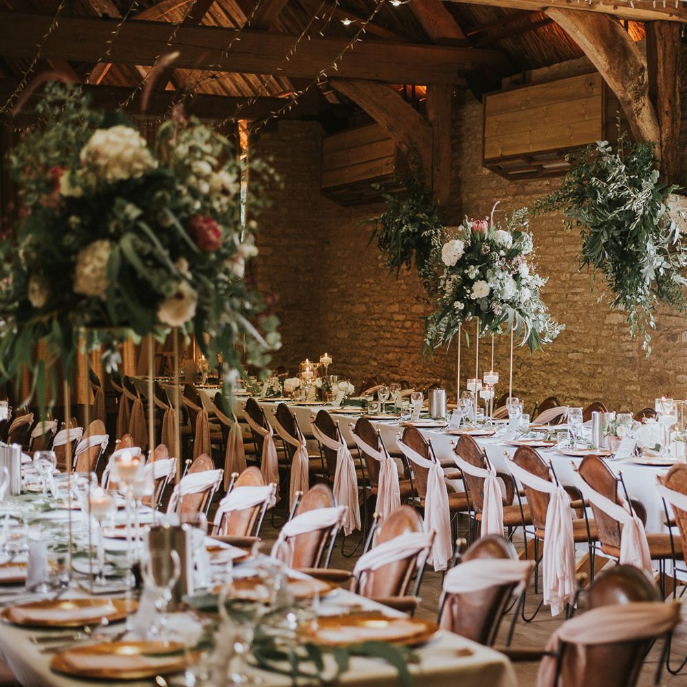 Brown leather chairs with blush pink fabric decor at long wooden breakfast tables with flower bombs and strung fairy lights inside stone room at Tythe Barn wedding with barn wedding flowers