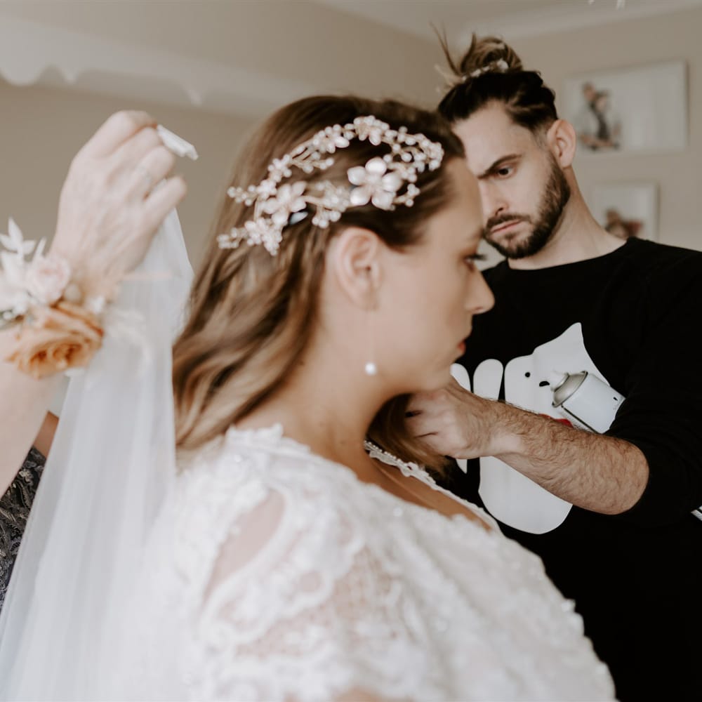 Bride in homemade lace wedding dress and floral and pearl hairpiece has her veil put on by wedding guest in blue dress before DIY garden wedding in Bedfordshire