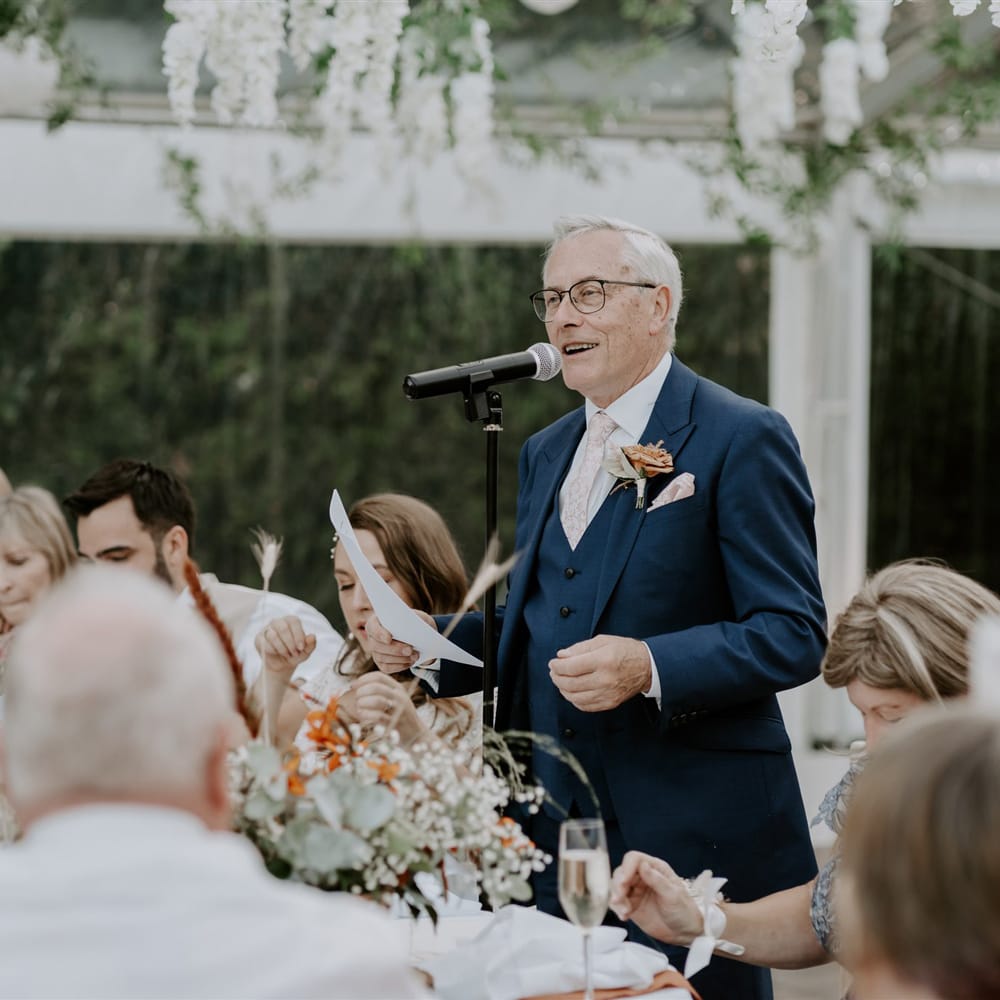 Man in blue three piece suit with burnt orange buttonhole makes speech during marquee wedding reception inn Bedfordshire 