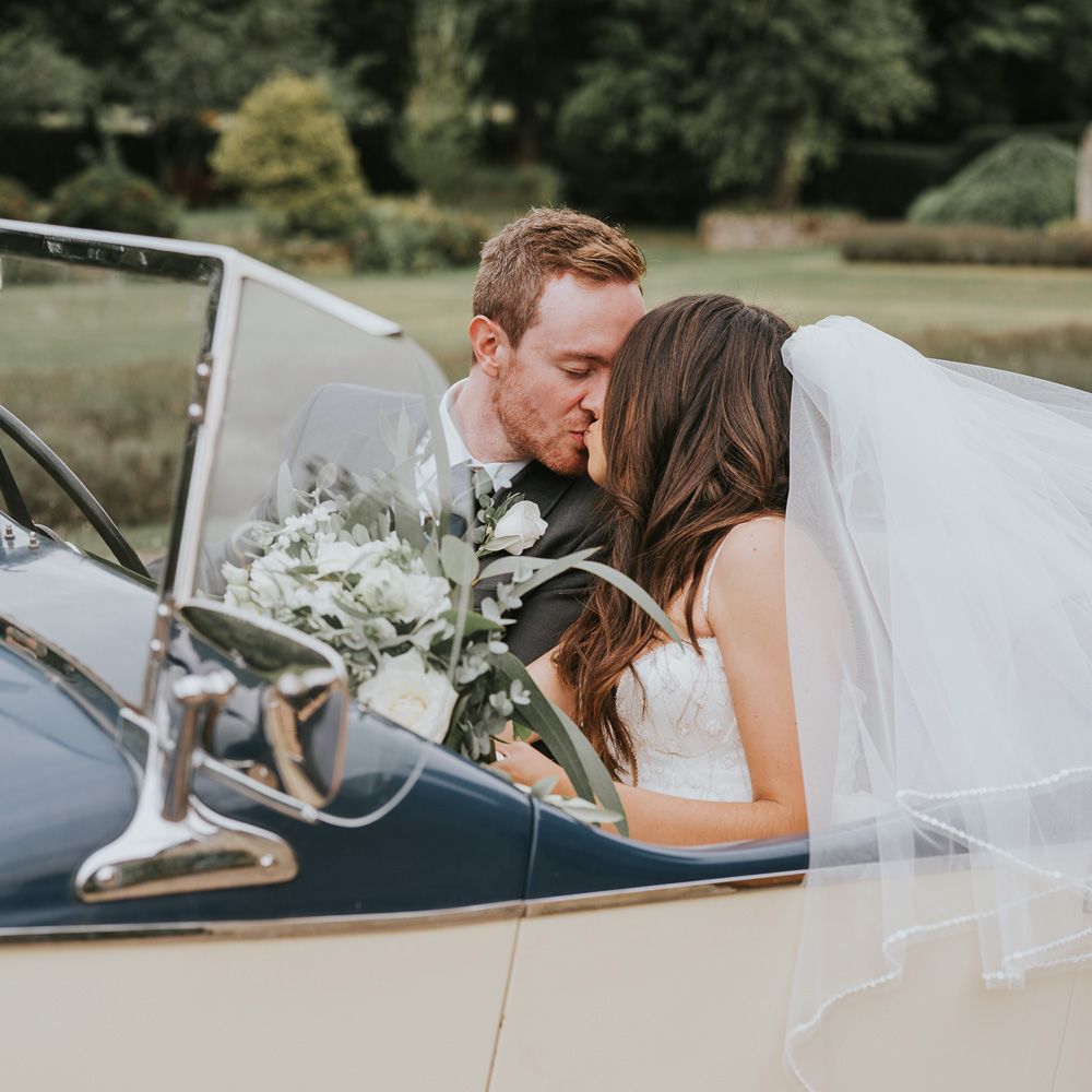 Bride in Pronovias wedding dress and veil holding white and green bridal bouquet kisses groom in grey suit as they sit in vintage car at Notley Abbey wedding