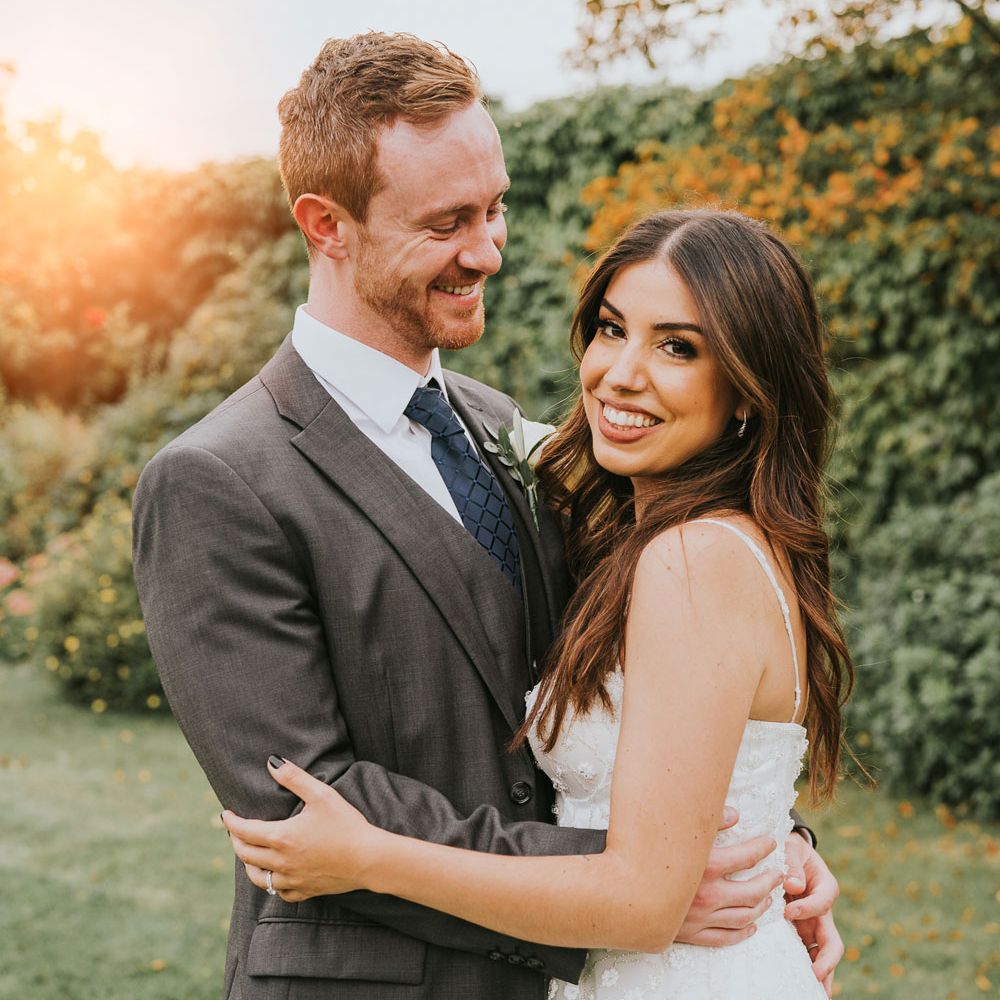 Groom in grey three piece suit, blue tie and white rose buttonhole smiles whilst looking at bride in white cami strap Pronovias wedding dress in the grounds of Notley Abbey in Buckinghamshire