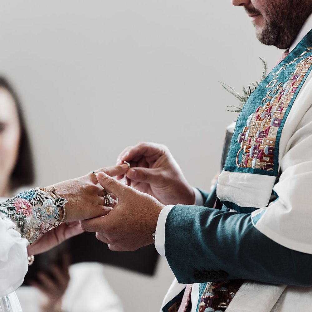 Groom in white and green tallit places ring on brides finger as she wears mesh sleeve wedding dress with embroidered cuff sleeves 