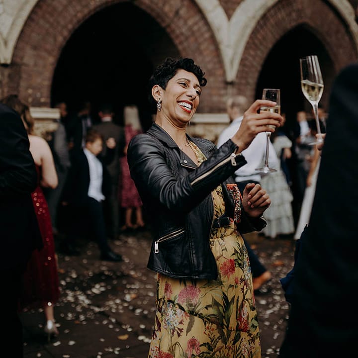 Wedding guest wears leather jacket and brightly coloured dress whilst hold champagne glass up to friend | Irene Yap Photography