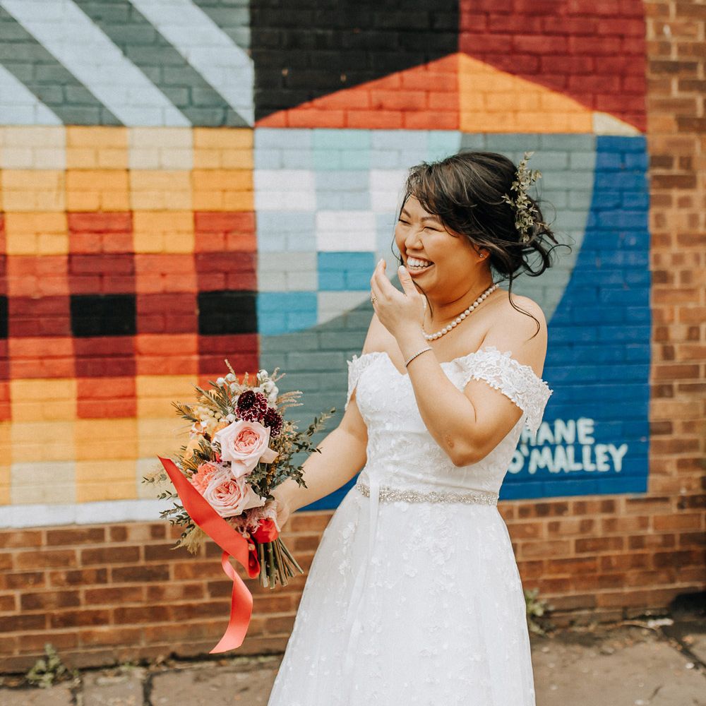 Laughing bride in wedding dress with belt holding mixed wedding bouquet with light pink roses as she stands by bright wall mural in Liverpool