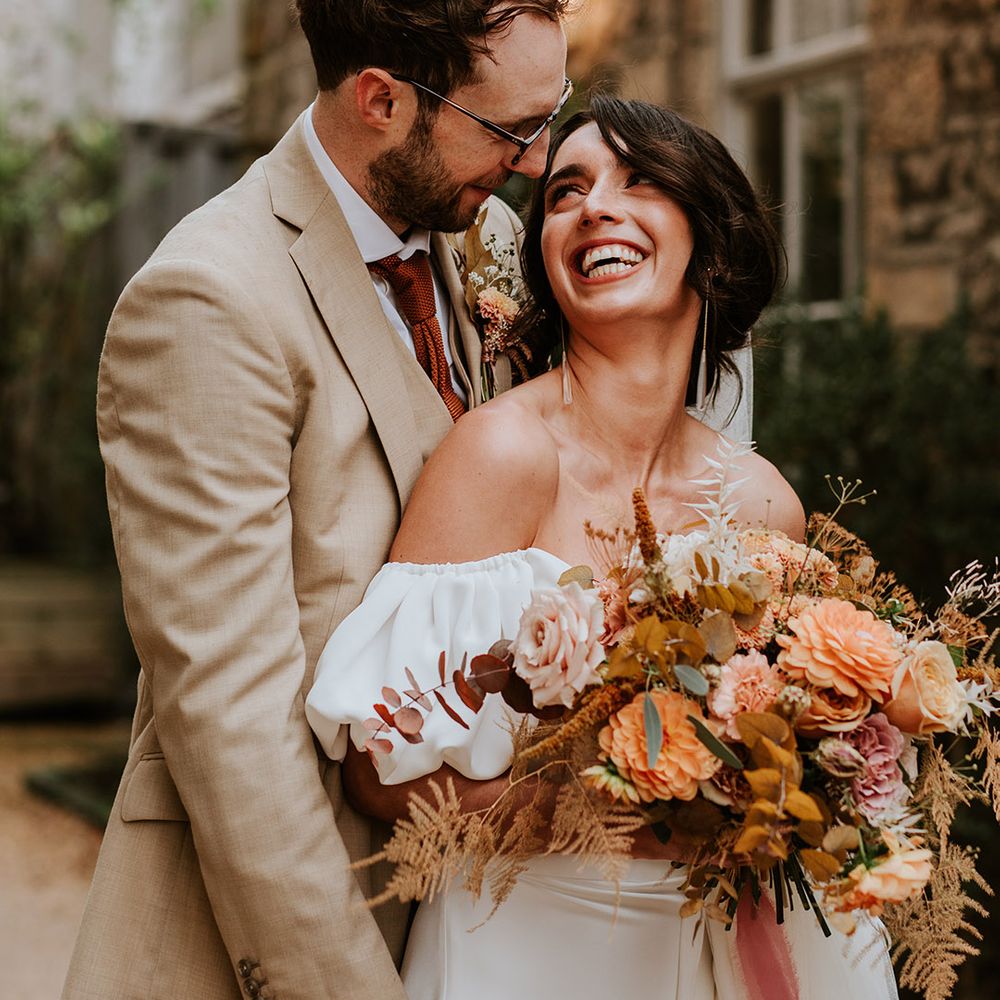 Groom in beige suit with orange tie hugs his bride form behind with bride holding autumnal wedding bouquet 