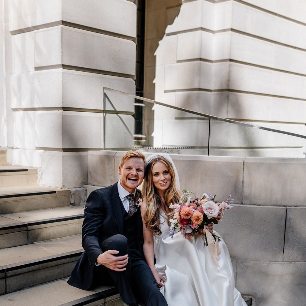 Bride in a princess wedding dress and long veil holding hands with her groom in a navy blue wool suit as they sit on the steps 