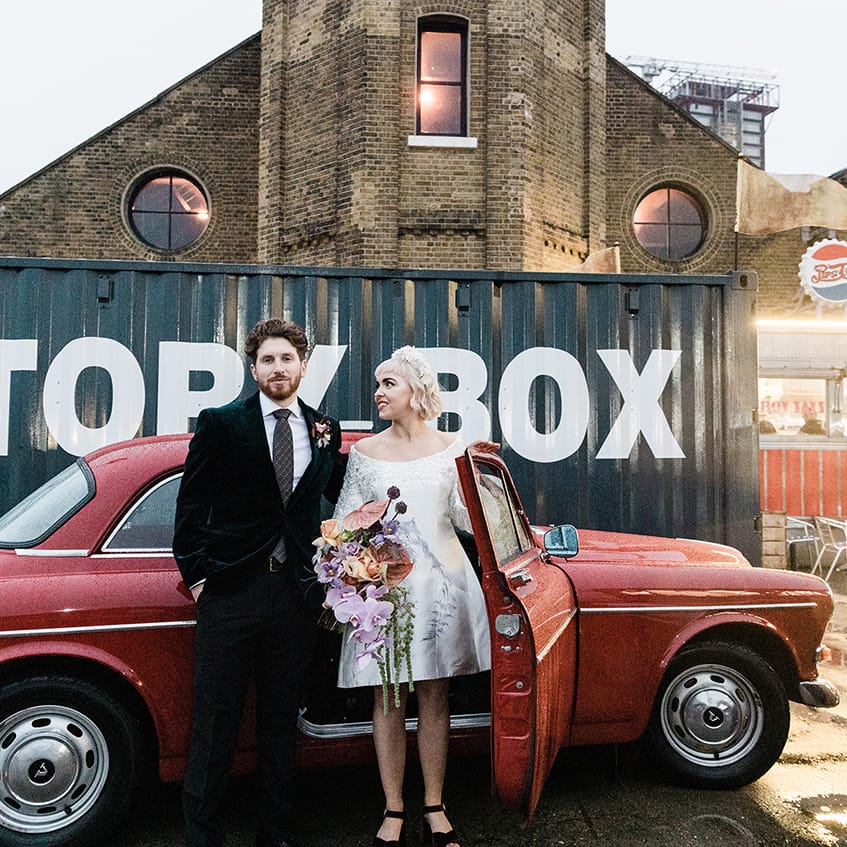 Bride and groom stand as they get out from their vintage red Volvo wedding car transport