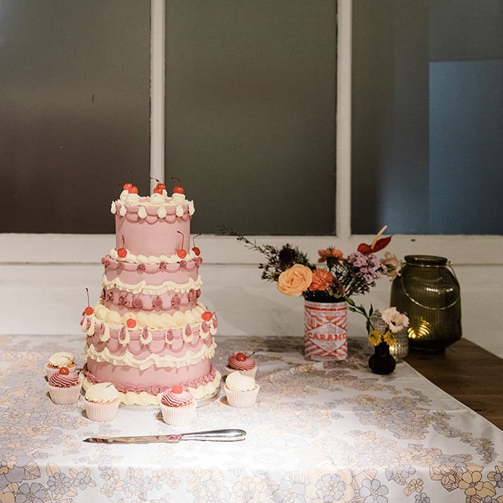 Three tier purple and white wedding cake with cherries and cupcakes in the same style as well as flowers in tin can and coloured glass vases