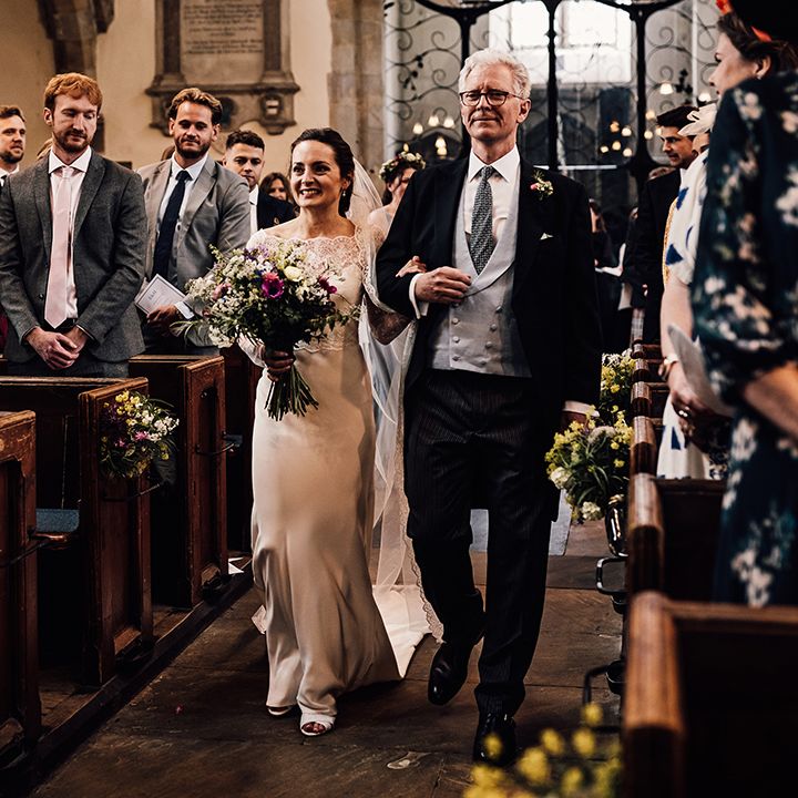 Father of the bride walks the bride in satin wedding dress with sheer lace sleeves down the aisle 