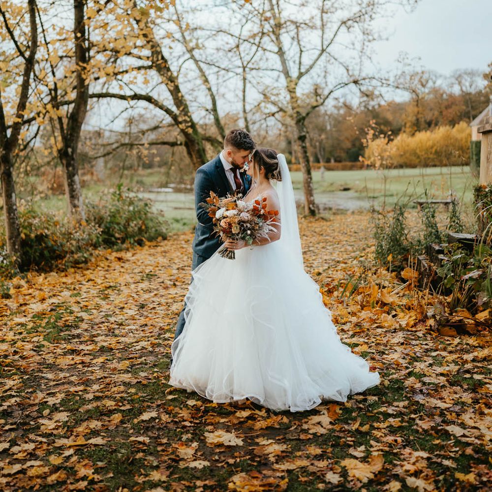 Bride and groom lean in close together as they stand in amongst autumn leaves 